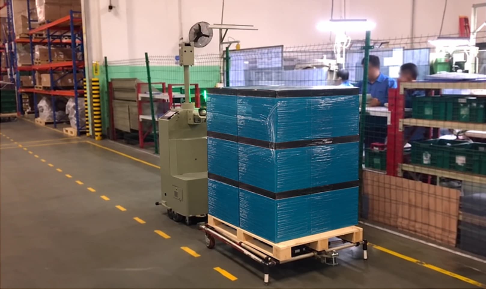 A tow AMR pulling a wrapped pallet on a dolly past a packing area